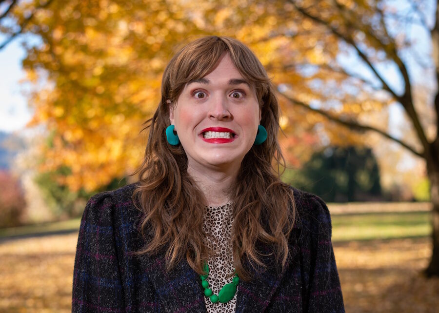 Portrait of Annabelle Wright with long wavy hair, teal earrings and a green necklace, wide-eyed with a big toothy grin in front of golden autumn trees.