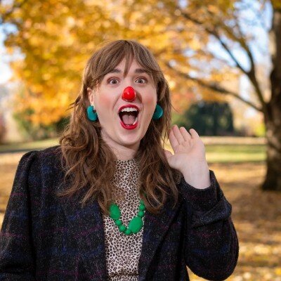 Annabelle Wright wearing a red clown nose, green earrings and necklace, hand cupped to her ear with a delighted open-mouthed expression in front of autumn foliage.